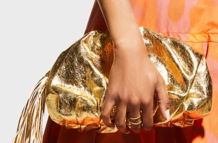 In "How to Dress for a Spring Wedding" by fashion stylist Jaquelyn Wahidi, a photo displaying a gold clutch against a woman wearing coral and orange dress is displayed.